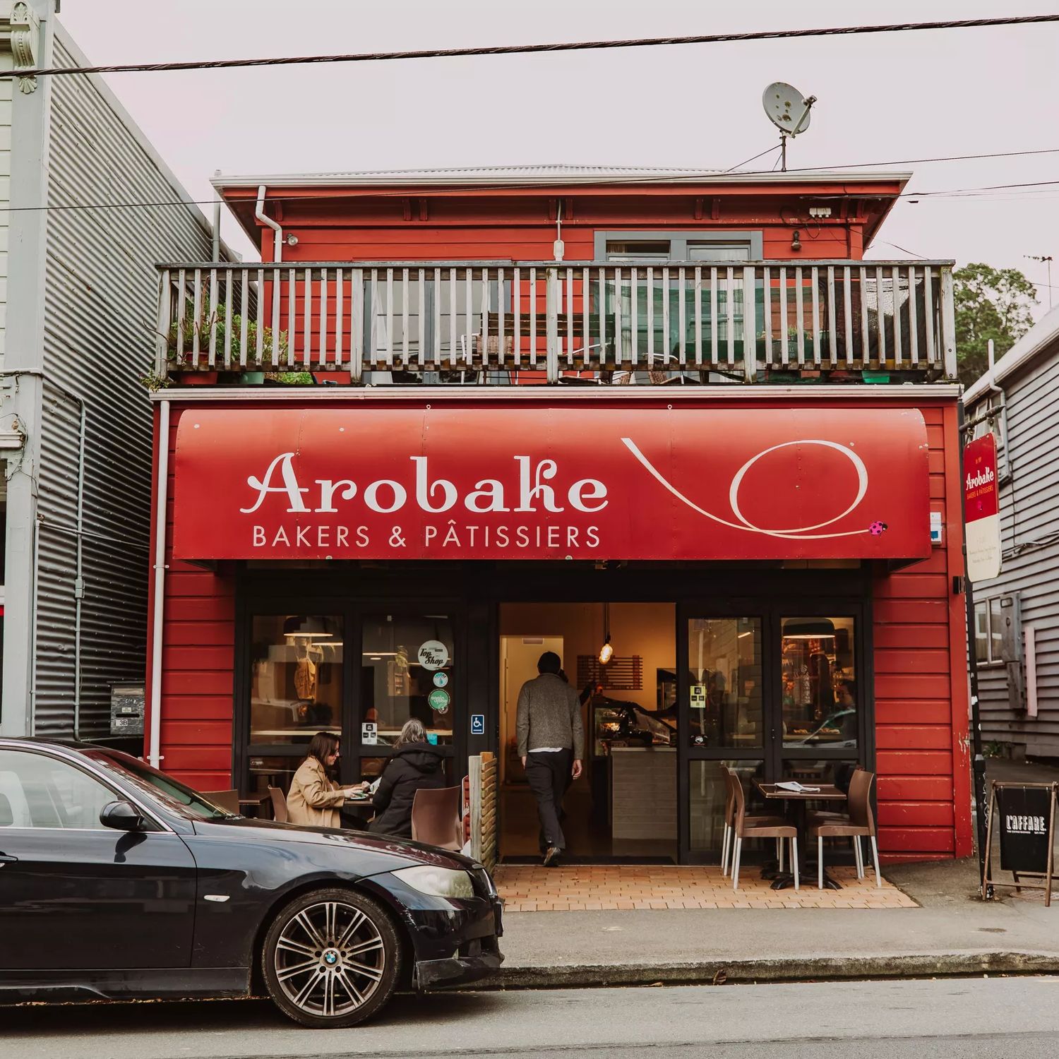 The front facade of Arobake Bakery in Aro Valley in Wellington. It is a small red-coloured two-story house with two four-seater tables outside.
