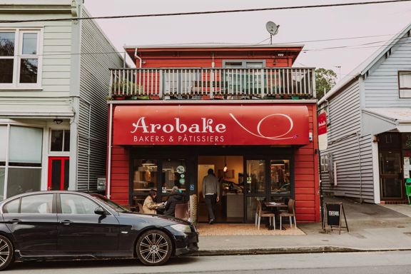 The front facade of Arobake Bakery in Aro Valley in Wellington. It is a small red-coloured two-story house with two four-seater tables outside.