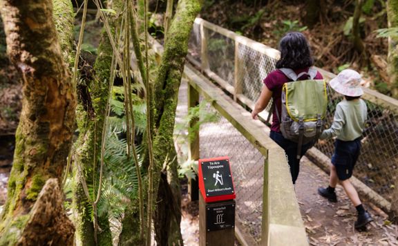 Adult and child walking on a bridge next to a red marker sign for Te Ara Tūpoupou.