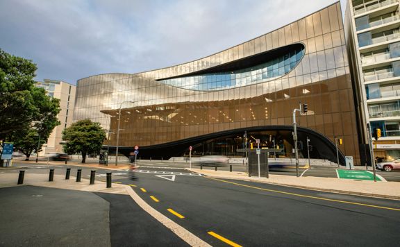 The exterior of Tākina from Cable street in day time, a tree sits on the left and the building itself has the reflection of blue skies.