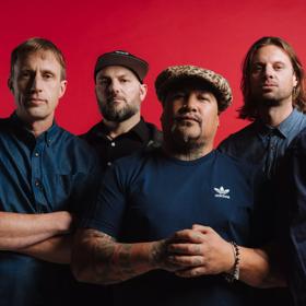 The five members of Shapeshifter, a live drum and bass act from New Zealand, wear navy blue and pose in front of a red wall.