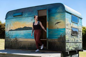 Person wearing activewear holding the door open as they walk out of a caravan. The caravan has a beach scene painted on it.