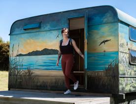 Person wearing activewear holding the door open as they walk out of a caravan. The caravan has a beach scene painted on it.