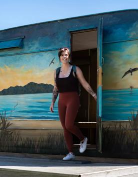 Person wearing activewear holding the door open as they walk out of a caravan. The caravan has a beach scene painted on it. 