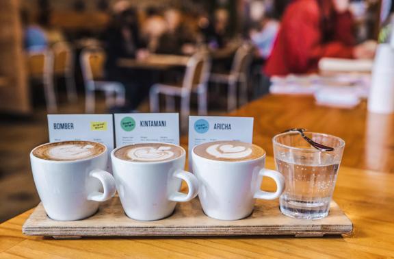 A flight of three artisanal coffees and a glass of water arranged neatly in a row placed on a wooden board on a table at The Hangar, a cafe in Te Aro Wellington. 