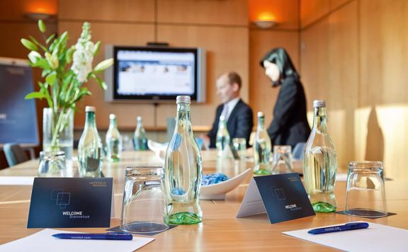 A meeting room setup at Novotel Wellington. There are two people at the table, the walls are paneled in a light-coloured wood and there are glass water bottles, drinking glasses pens and paper prepared at each seat.