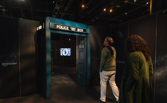 Two people approaching an archway that resembles the Doctor Who Tardis. The door way is navy blue and has a light up sign on top that reads Police Public Call Box.