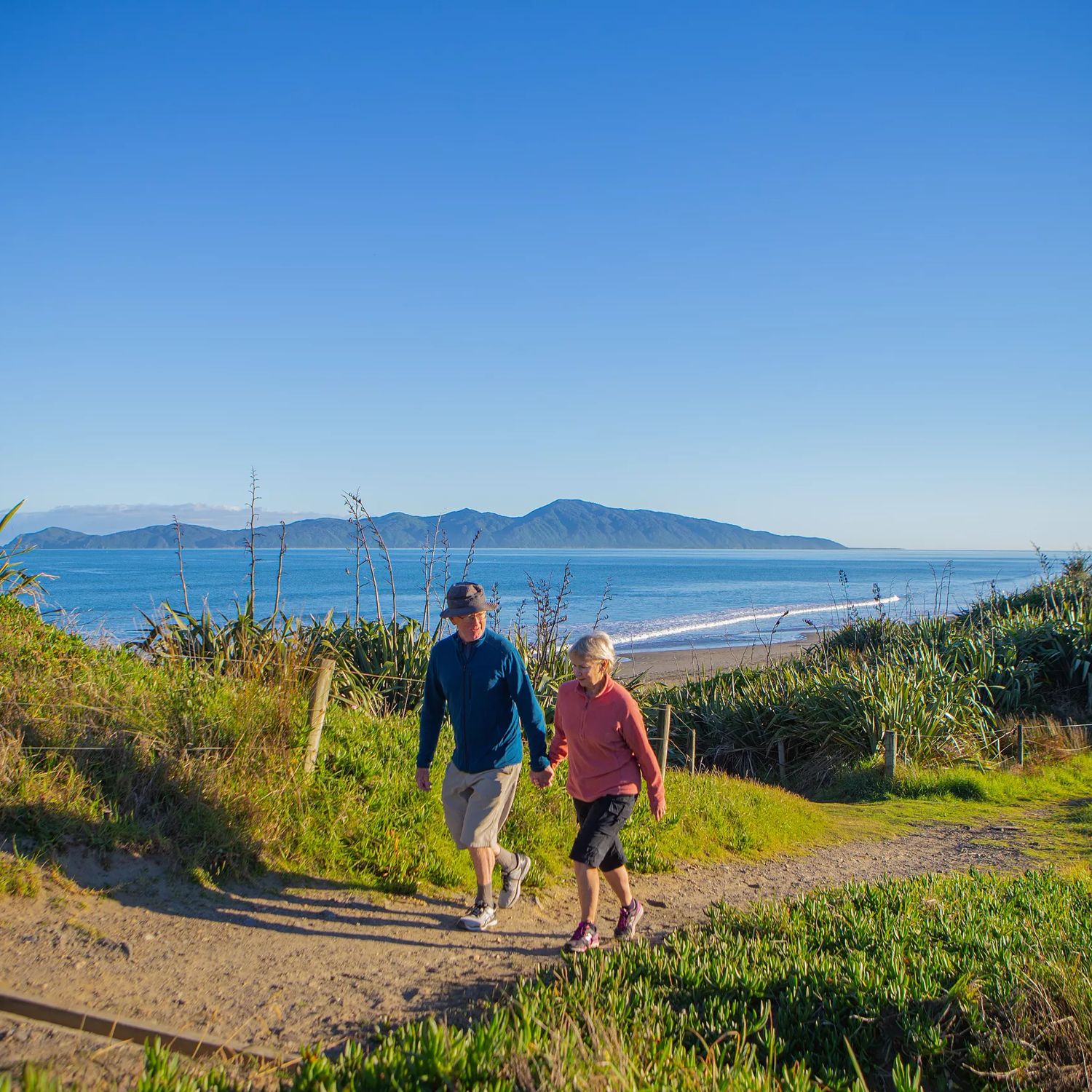 Two people hold hands and walk on a path in the Queen Elizabeth Park Regional Park located on the Kāpiti Coast in New Zealand. The ocean and Kapiti Island are visible in the background.