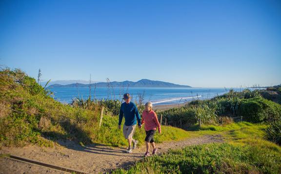 Two people hold hands and walk on a path in the Queen Elizabeth Park Regional Park located on the Kāpiti Coast in New Zealand. The ocean and Kapiti Island are visible in the background.