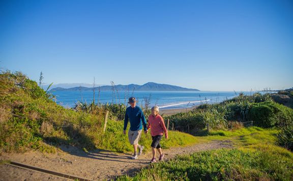 Two people hold hands and walk on a path in the Queen Elizabeth Park Regional Park located on the Kāpiti Coast in New Zealand. The ocean and Kapiti Island are visible in the background.