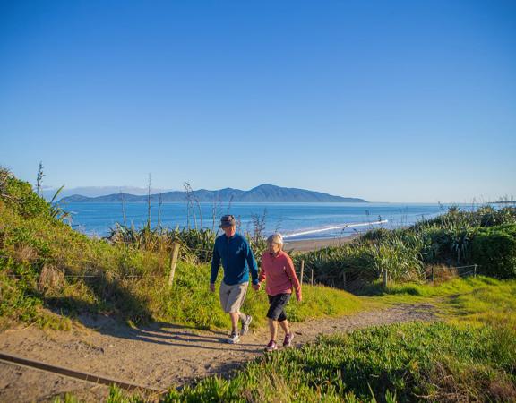 Two people hold hands and walk on a path in the Queen Elizabeth Park Regional Park located on the Kāpiti Coast in New Zealand. The ocean and Kapiti Island are visible in the background.