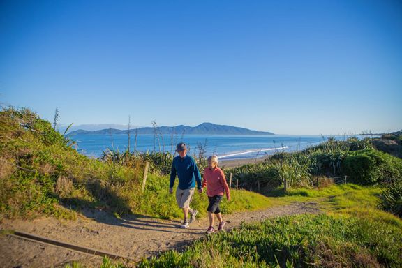 Two people hold hands and walk on a path in the Queen Elizabeth Park Regional Park located on the Kāpiti Coast in New Zealand. The ocean and Kapiti Island are visible in the background.