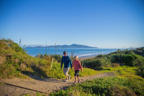 Two people hold hands and walk on a path in the Queen Elizabeth Park Regional Park located on the Kāpiti Coast in New Zealand. The ocean and Kapiti Island are visible in the background. 