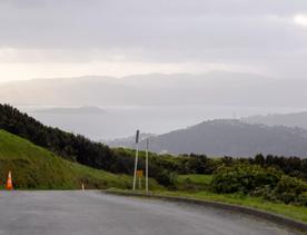 The Brooklyn Wind Turbine sits on a hill above Wellington, with views of the city. Bush and trees surround the area.