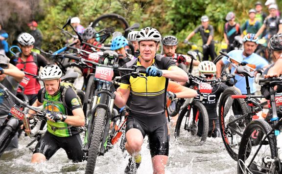 A mass of mountain bikers in lycra and helmets carry their bikes as they cross a river.