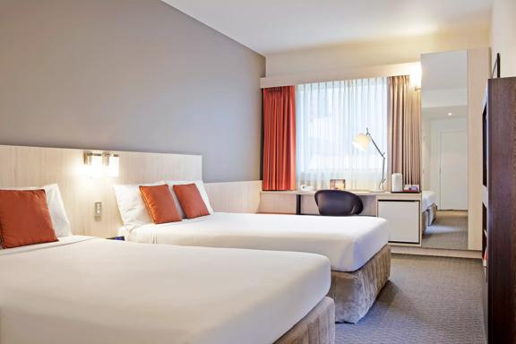 A room at the Ibis Wellington. It has two queen beds with perfectly smooth white bedding each with two orange pillows. A desk and chair sit against the window and a full-length mirror off to the side reflects the room.