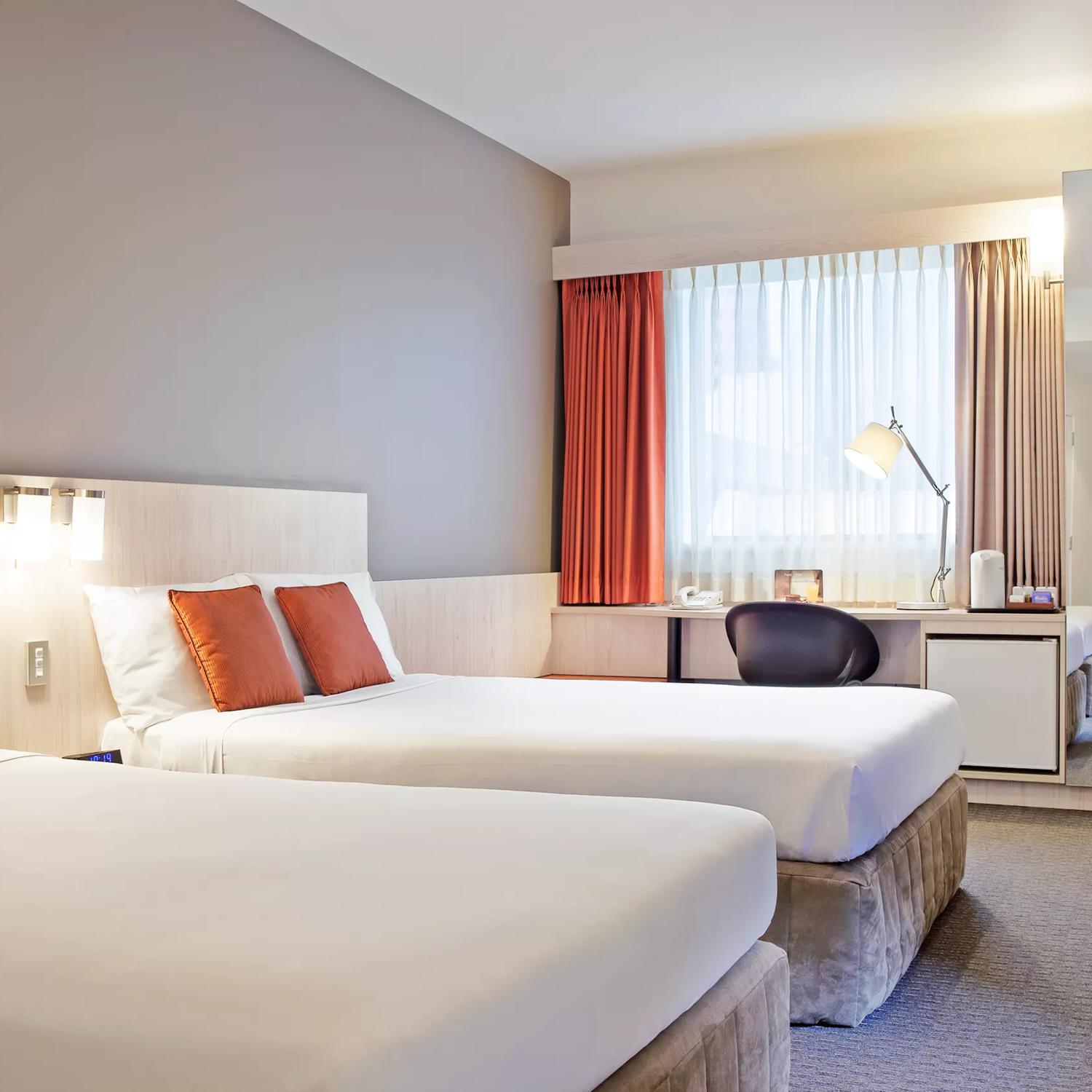 A room at the Ibis Wellington. It has two queen beds with perfectly smooth white bedding each with two orange pillows. A desk and chair sit against the window and a full-length mirror off to the side reflects the room.