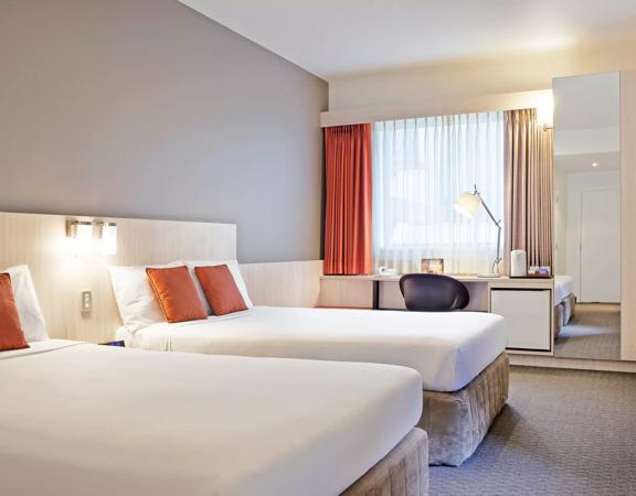 A room at the Ibis Wellington. It has two queen beds with perfectly smooth white bedding each with two orange pillows. A desk and chair sit against the window and a full-length mirror off to the side reflects the room.