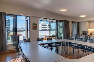 Interior of one of AREA's conference rooms, depicting tables configured into a square in the foreground, with a kitchen and floor-to-ceiling windows in background and balcony that has views of Wellington Central buildings.