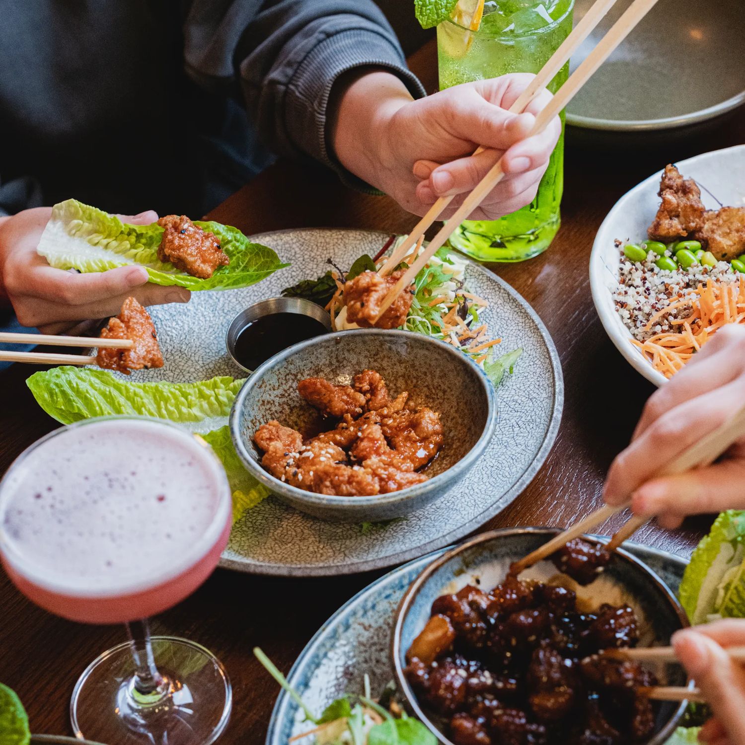 Plates of food, including Asian style proteins and accoutrements with hands using chopsticks and a couple of colourful cocktails.