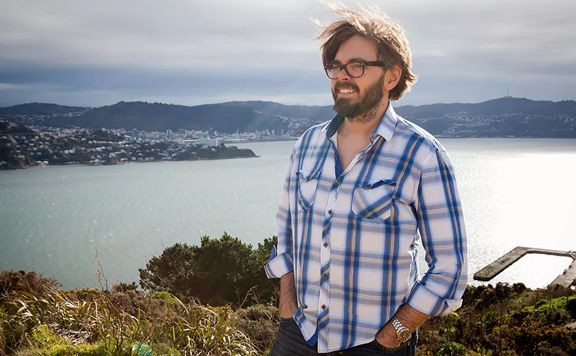 Series director of the TV show ‘Thunderbirds Are Go’ David Scott stands on top of Miramar Peninsula with Wellington city in the background. He wears a blue and white check shirt and has wind-swept hair.