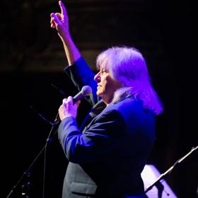 Rodger Fox performs on stage under a purple spotlight with one arm raised in the air and the other holding a microphone.