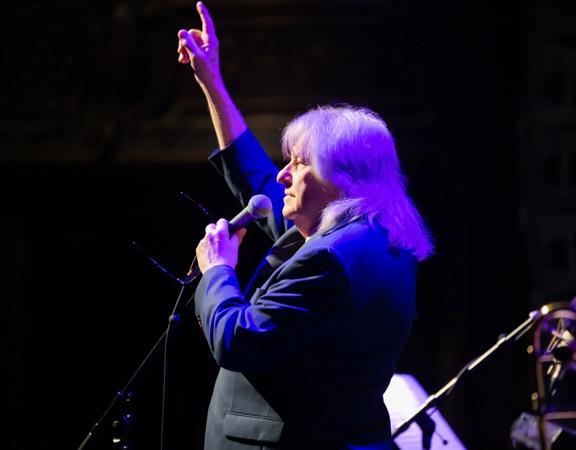 Rodger Fox performs on stage under a purple spotlight with one arm raised in the air and the other holding a microphone.