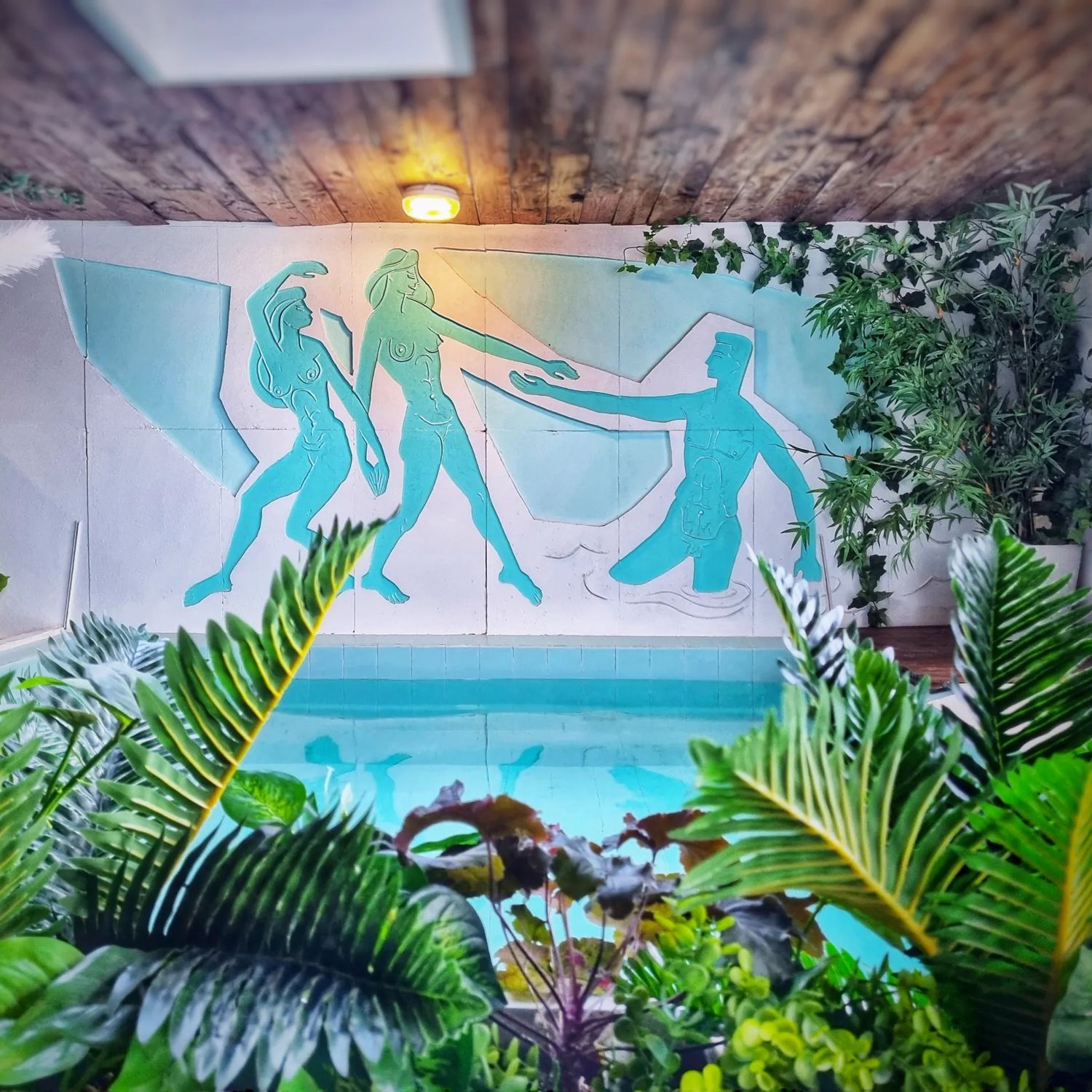 A blue mural wall above a pool inside Tory Urban Retreat, depicting nude people. Artificial plants surround the room.