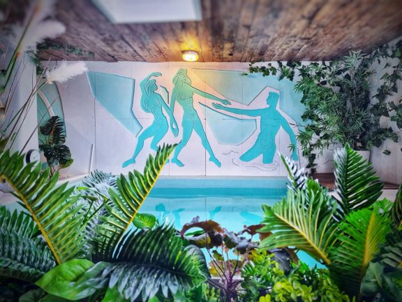 A blue mural wall above a pool inside Tory Urban Retreat, depicting nude people. Artificial plants surround the room.