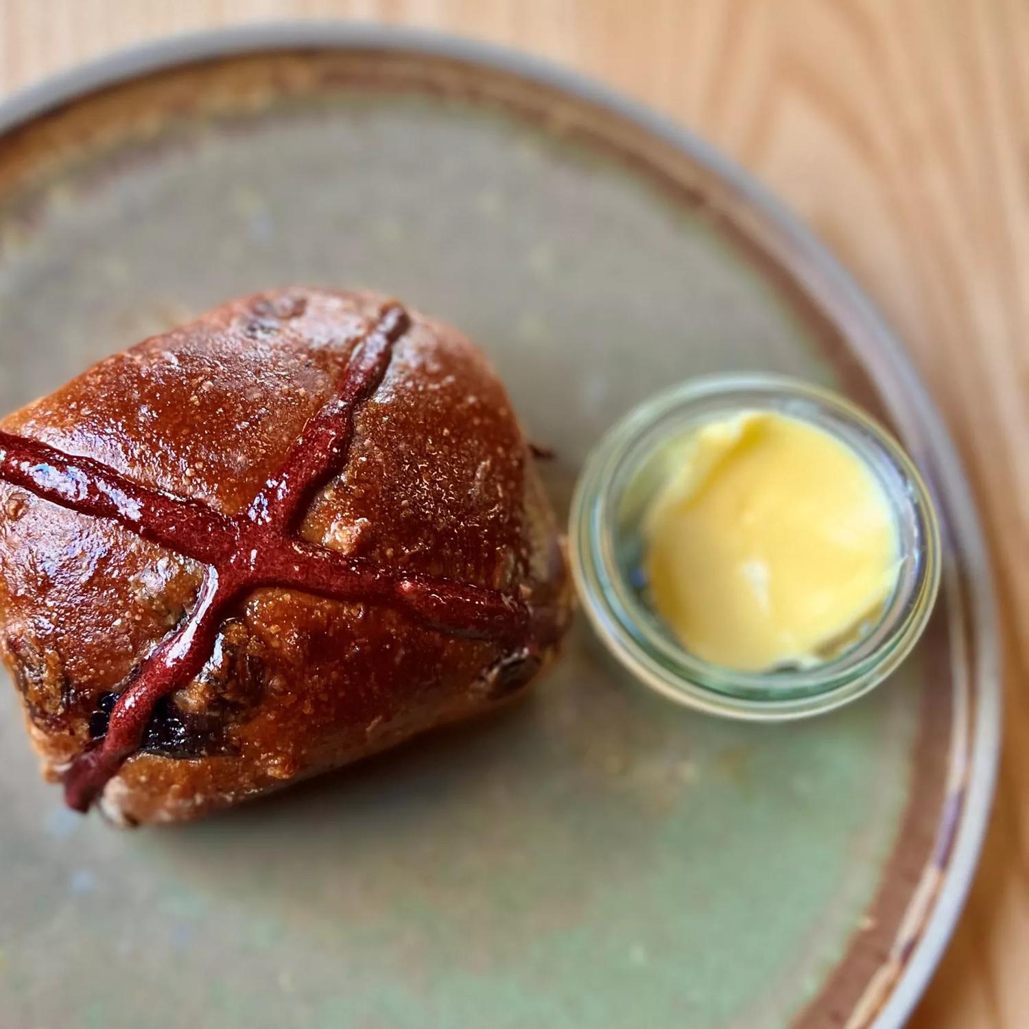 A single hot cross bun from August café. It is pictured looking directly down from above. It sits on a handmade pottery plate with a small bowl of butter to the right hand side.