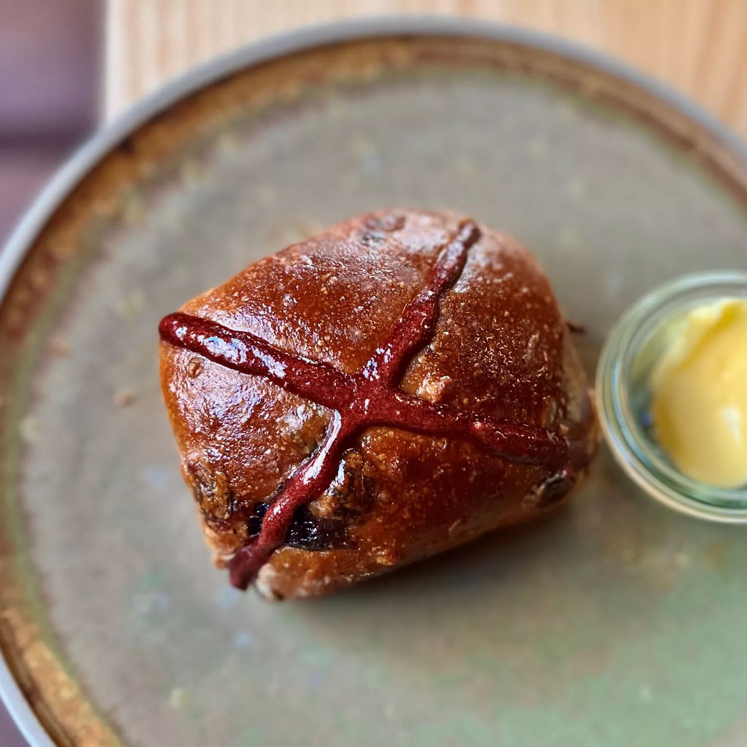 A single hot cross bun from August café. It is pictured looking directly down from above. It sits on a handmade pottery plate with a small bowl of butter to the right hand side.