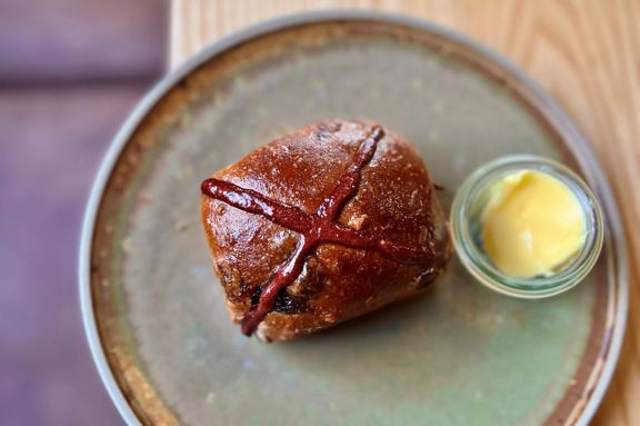 A single hot cross bun from August café. It is pictured looking directly down from above. It sits on a handmade pottery plate with a small bowl of butter to the right hand side.