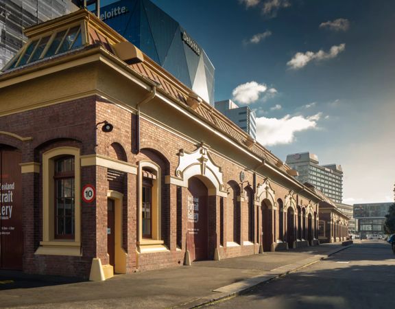 The exterior of the New Zealand Portrait Gallery — a historic brick shed on the waterfront, with city buildings behind it.