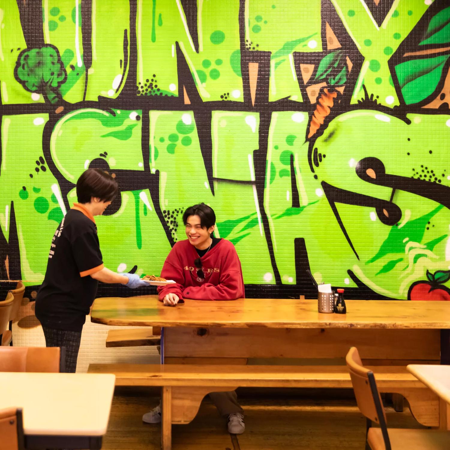 Inside Aunty Mena's Vegetarian Restaurant & Café, a popular Malaysian restaurant on Cuba Street in Te Aro, Wellington. A worker wearing black hands a dish to a smiling customer at a large wooden table and the restaurant's name is graffitied in big lime gr