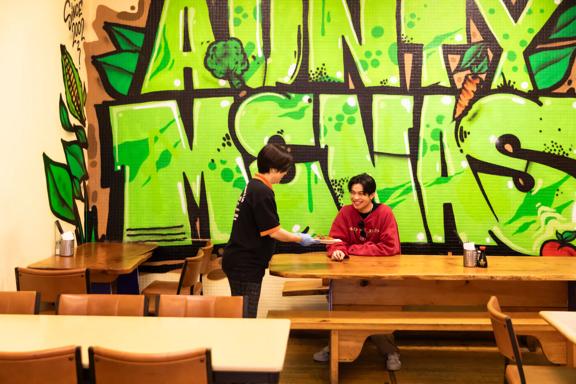 Inside Aunty Mena's Vegetarian Restaurant & Café, a popular Malaysian restaurant on Cuba Street in Te Aro, Wellington. A worker wearing black hands a dish to a smiling customer at a large wooden table and the restaurant's name is graffitied in big lime gr
