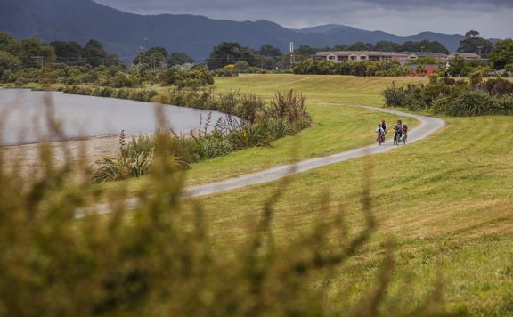 Two adults and two children riding bicycles on a gravel path surrounded by green grass, flax, and the Hutt River flowing nearby.