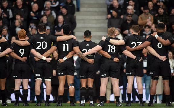 The All Blacks rugby team stands arm-in-arm facing the crowd in the stadium before a match. 