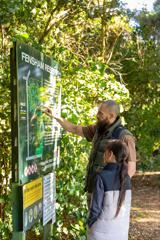 Two people standing at an information board at Fensham Reserve in Wairarapa, surrounded by lush green foliage. One person is pointing to a map of walking tracks.