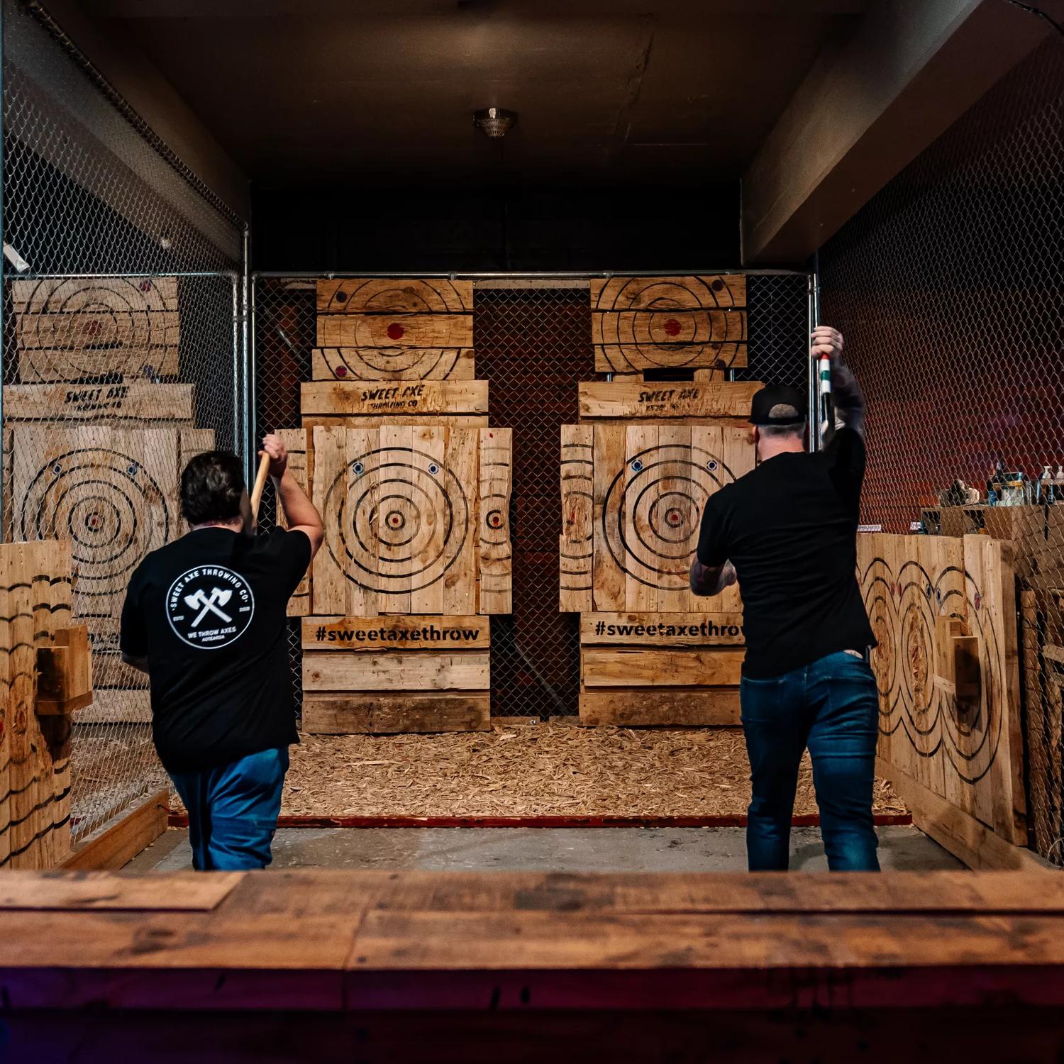 Two people prepare to throw axes at wooden targets at Sweet Axe Throwing. Metal fences line the throwing area.