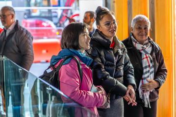 Three people wearing jackets stand beside one another looking at something out of frame. There are two other people and a large glass window in the background.