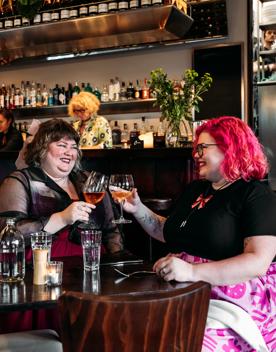 Two people enjoying a meal and clinking their wine glasses in Capitol Restaurant. Waitstaff make drinks behind the bar behind them. The dark wood tables add a moody vibe to the interior.