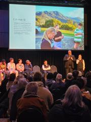 An adult and a child on stage speaking to a large group of seated conference delegates. They are joined by five people on stage.