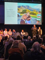 An adult and a child on stage speaking to a large group of seated conference delegates. They are joined by five people on stage.