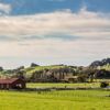 A dark red farmhouse surrounded by green farmland and rolling hills in the background.
