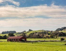 A dark red farmhouse surrounded by green farmland and rolling hills in the background.
