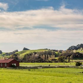 A dark red farmhouse surrounded by green farmland and rolling hills in the background.
