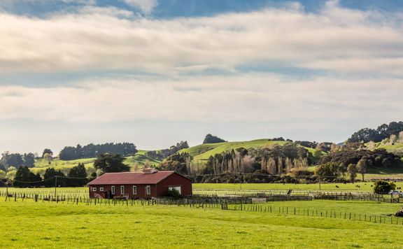 A dark red farmhouse surrounded by green farmland and rolling hills in the background.