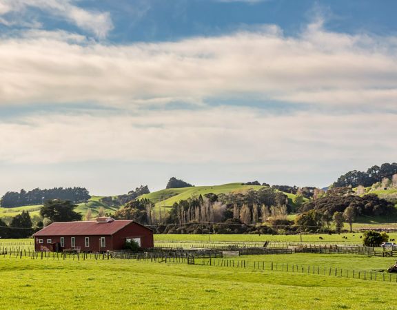 A dark red farmhouse surrounded by green farmland and rolling hills in the background.