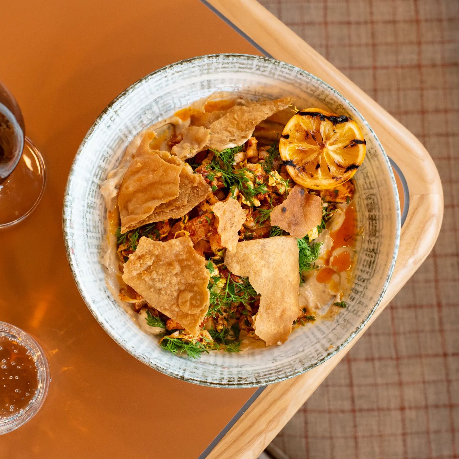 A plate of hummus garnished with fresh herbs, a grilled lemon, and pita chips, a glass of red wine and a glass of sparkling water at Parla, a Mediterranean restaurant in Island Bay, Wellington.