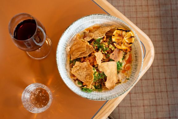 A plate of hummus garnished with fresh herbs, a grilled lemon, and pita chips, a glass of red wine and a glass of sparkling water at Parla, a Mediterranean restaurant in Island Bay, Wellington.